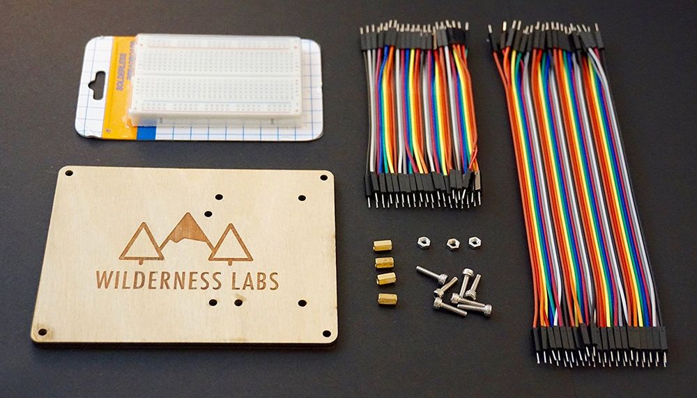 Photo of a Wilderness Labs hack kit showing a breadboard, branded wooden mounting board, several flexible wires with pin headers, and mounting hardware.